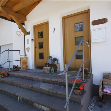 An inviting house entrance with a wooden staircase and two doors. Colorful flowers and a bench in the entrance area create a friendly impression.