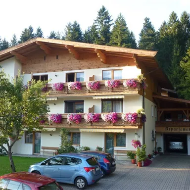 A charming multi-story house surrounded by trees. The balcony is adorned with colorful flowers, and cars are parked in the foreground.