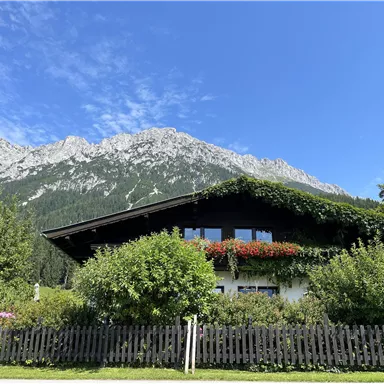 A charming house surrounded by lush greenery and colorful flowers. In the background, majestic mountains rise under a clear blue sky.
