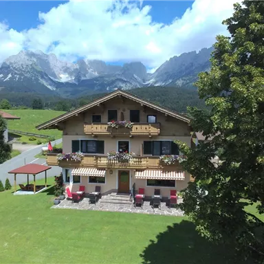 Ein gemütliches Haus in den Bergen mit blühenden Balkonen. Im Hintergrund sind majestätische Berge und ein klarer blauer Himmel zu sehen.