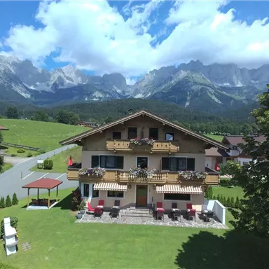 Ein schönes Chalet mit Balkonen und Blumen. Im Hintergrund sind beeindruckende Berge und ein blauer Himmel zu sehen.