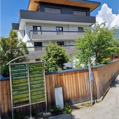 A modern building with several balconies and a green environment. On the left side, there is an information sign displaying various offers.