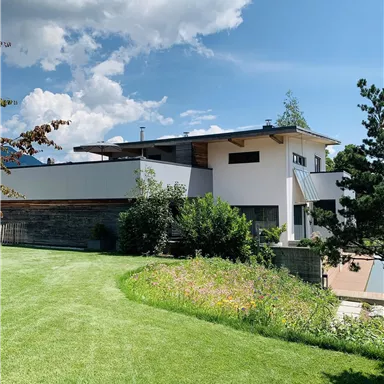 A modern house with a green lawn and colorful flowers in the foreground. The sky is blue with some clouds.