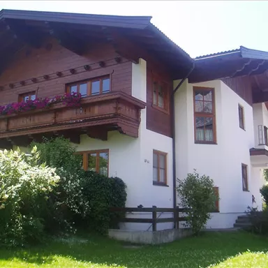 Ein schönes Haus im alpenländischen Stil mit einem Holzbalkon und blühenden Blumen. Grüne Sträucher umgeben das Gebäude und der Himmel ist klar.