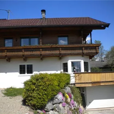 Ein schönes Haus im alpinen Stil mit einem Holzbalcony. Der Garten ist mit bunten Blumen geschmückt und der Himmel ist klar und blau.