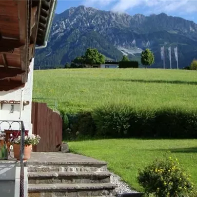 Ein malerischer Blick auf eine grüne Wiese mit Bergen im Hintergrund. Im Vordergrund sieht man eine Terrasse mit Stühlen und einen kleinen Garten.