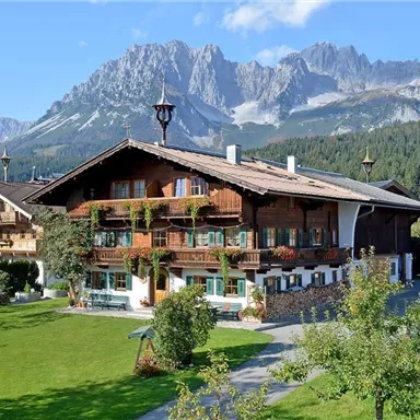 Ein charmantes alpenländisches Haus mit Holzfassade und blühenden Balkonen. Im Hintergrund erheben sich majestätische Berge unter einem blauen Himmel.