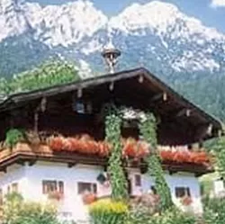 Ein traditionelles Alpenhaus mit blühenden Balkonen. Im Hintergrund erheben sich majestätische Berge unter einem klaren Himmel.