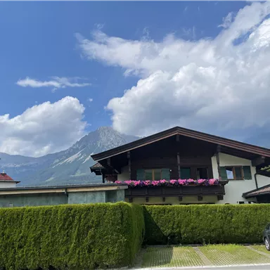 Ein schönes Haus mit einem Garten und bunten Blumen auf dem Balkon. Im Hintergrund sind Berge und ein blauer Himmel zu sehen.