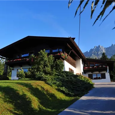 Ein schönes Haus umgeben von Grün und mit blühenden Fenstern. Im Hintergrund sind die beeindruckenden Berge zu sehen.