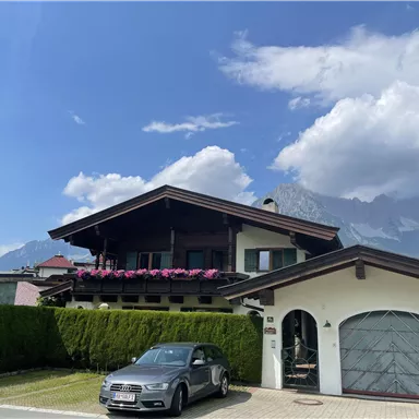 Ein schönes Haus mit Balkonen und bunten Blumen. Im Hintergrund sind Berge und ein klarer Himmel zu sehen.