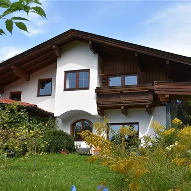 Ein charmantes Haus mit Holzverkleidung und großen Fenstern in einem grünen Garten. Im Hintergrund sind sanfte Hügel sichtbar.