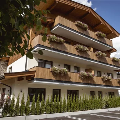 Ein modernes Gebäude mit holzverkleideten Balkonen und vielen Blumen. Es befindet sich in einer ruhigen Umgebung mit Bergen im Hintergrund.