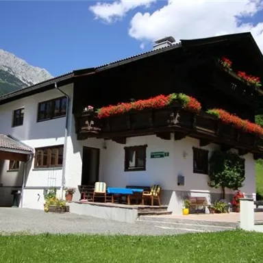Ein gemütliches Haus im alpinen Stil mit einem Balkon voller Blumen. Im Hintergrund sind grüne Wiesen und Berge sichtbar.