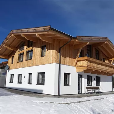 A modern chalet with wooden cladding and a white paint. The snow covers the ground and the view of the mountains is clear.