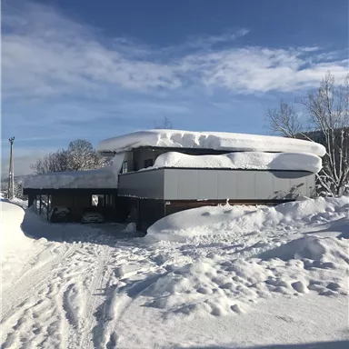 A modern house amidst deep snow. The sky is clear and blue, while a thick layer of snow rests on the roof.