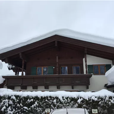 Ein chaletartiges Haus mit einem schneebedeckten Dach und großen Fenstern. Der Bereich vor dem Haus ist ebenfalls mit Schnee bedeckt.