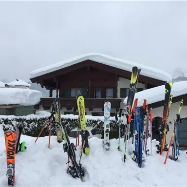 Ein Chalet im Schnee mit vielen Skiern vor der Tür. Der Himmel ist bewölkt und die Umgebung ist winterlich.