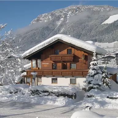 Ein gemütliches Holzhaus im Schnee, umgeben von verschneiten Bäumen. Im Hintergrund erheben sich majestätische Berge unter einem klaren blauen Himmel.
