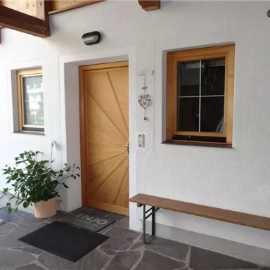 Ein einladender Eingangsbereich mit einer Holztür und zwei Fenstern. Neben der Tür steht eine grüne Pflanze in einem Topf und eine Bank ist davor platziert.