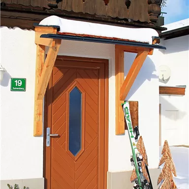 Eine gemütliche Haustür mit Holzverkleidung in einer schneebedeckten Landschaft. Skier stehen neben der Tür, und der Himmel ist blau.