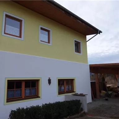 Ein modernes zweistöckiges Haus mit einer gelben und weißen Fassade. Im Vordergrund sind Fenster mit Holzrahmen und ein angrenzender überdachter Bereich sichtbar.