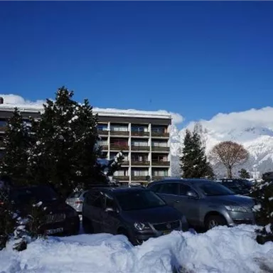 A building surrounded by snow, with a clear blue sky in the background. Parked cars and trees are also visible.