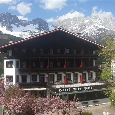 Ein traditionelles Alpenhotel mit einem blühenden Garten im Vordergrund. Im Hintergrund sind majestätische Berge und ein strahlend blauer Himmel zu sehen.