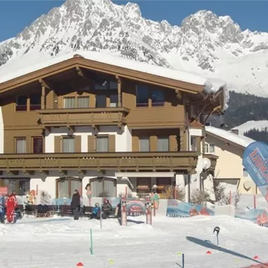 Ein gemütliches Chalet in den Bergen, umgeben von Schnee. Im Vordergrund sind Menschen, die Ski fahren und sich entspannen.