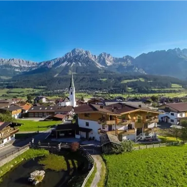 Eine malerische Dorfansicht mit traditionellen Häusern und einer Kirche, umgeben von grünen Wiesen und Bergen. Der Himmel ist klar und blau, was die idyllische Landschaft betont.