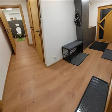 A bright hallway with a wooden floor and various doors. A picture of a forest landscape hangs on the wall.