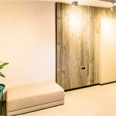 A modern entrance area with a padded stool and a plant on a table. The walls are in neutral tones, with wood paneling.