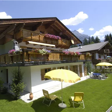 Ein gemütliches Haus im alpinen Stil mit Balkon und bunten Blumen. Im Vordergrund befindet sich ein gepflegter Garten mit sonnigen Liegen und Sonnenschirmen.