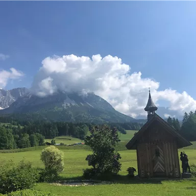 Eine malerische Landschaft mit Bergen im Hintergrund und einer kleinen Holzkirche im Vordergrund. Der Himmel ist blau mit einigen Wolken, und die Wiese ist grün und üppig.