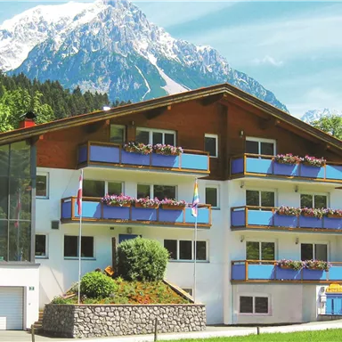 Ein modernes Haus mit mehreren Balkonen, umgeben von grünen Bäumen. Im Hintergrund erheben sich majestätische, schneebedeckte Berge.