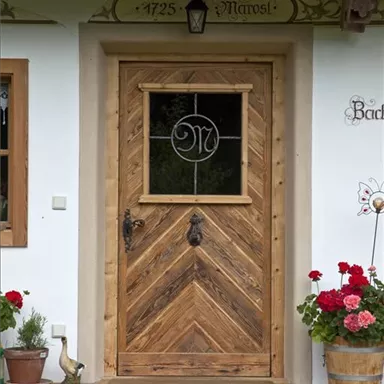 Eine rustikale Holztür mit einem eleganten Fenster in der Mitte. Vor der Tür blühen rote Geranien und ein kleines Dekorationsobjekt steht im Garten.