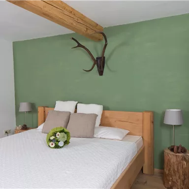 A cozy bedroom with a large bed and a green accent wall. Decorative elements like a flower and a deer antler add rustic charm to the space.