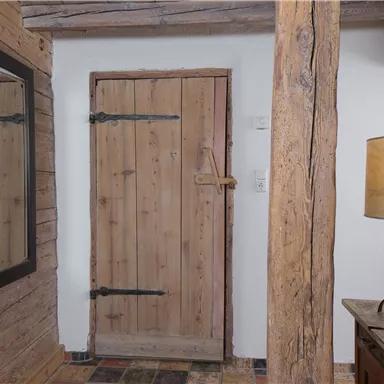 A rustic entrance area with a wooden-clad door and a large mirror. Next to the door stands an elegant lamp on a wooden sideboard.