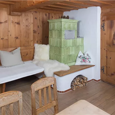 A cozy room with wood paneling and a green tiled stove. A comfortable seating area and a wooden table give the room an inviting atmosphere.