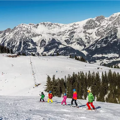 Eine Gruppe von Kindern fährt Ski auf einem schneebedeckten Hang. Im Hintergrund sind beeindruckende Berge und ein klarer blauer Himmel zu sehen.