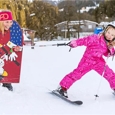 Ein fröhliches Kind in einem pinken Skianzug fährt Ski auf verschneitem Boden. Im Hintergrund steht ein lustiges Spiel mit einer Figur und einem Clown.
