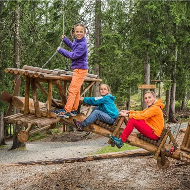 Drei Kinder spielen auf einem Holzwagen im Wald. Sie haben Freude und tragen bunte Kleidung.