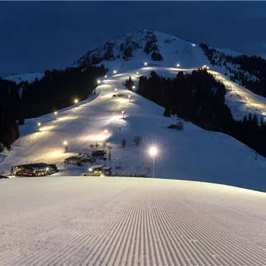 Eine verschneite Berglandschaft bei Nacht, beleuchtet von vielen Lichtern. Im Vordergrund ist die präparierte Piste sichtbar.