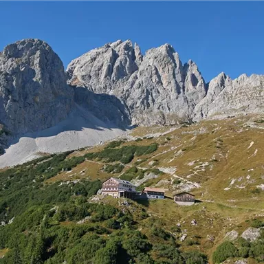An impressive mountain landscape with steep rocks and green terrain. In the middle, there is a mountain inn, surrounded by nature.