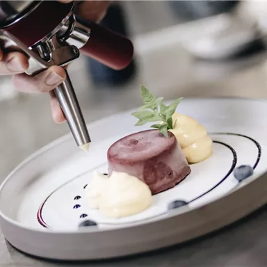 Eine Person verziert ein Dessert auf einem weißen Teller. Das Dessert besteht aus einem roten Gelee und zwei Häufchen Sahne, dekoriert mit einem Blatt Minze.