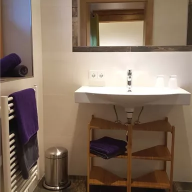 A modern bathroom with a sink, mirror, and towels. The floor is made of dark tiles, and there is a wooden shelf for additional towels.