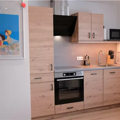 Eine moderne Küche mit Holzschränken, einem Herd und einer Mikrowelle. An der Wand hängt ein farbenfrohes Bild von "Heidi".