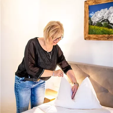 Eine Frau richtet das Kopfkissen auf einem Bett. Im Hintergrund ist ein Bild von Bergen zu sehen.