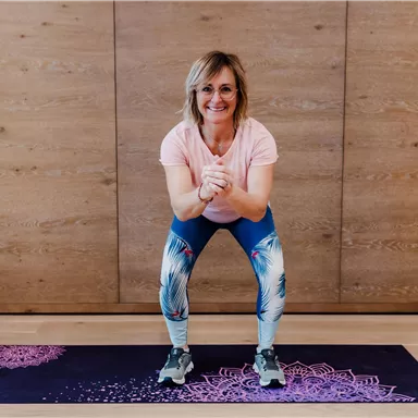 A woman is doing squats on a yoga mat. She is wearing colorful leggings and smiling.