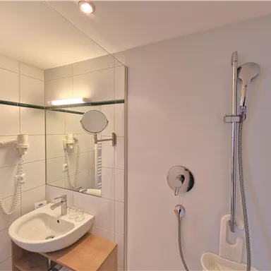 Ein modernes Badezimmer mit einer Dusche und einem Waschbecken. Die Wände sind mit weißen Fliesen gefliest und es gibt einen Spiegel.